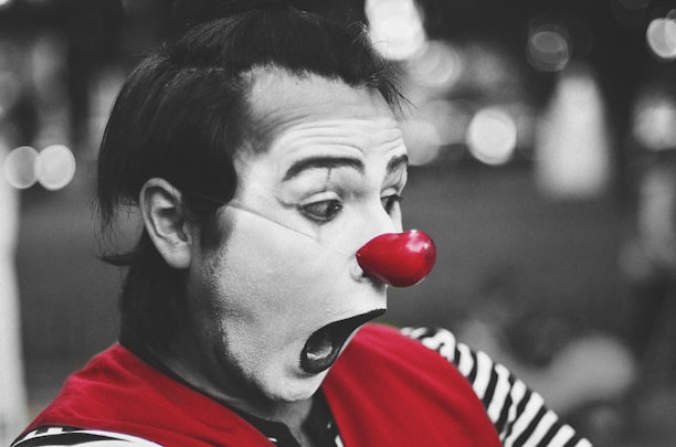 A clown with white face paint and exaggerated facial features is wearing a bright red nose and a red garment over a black and white striped shirt. The focus is on the clown's expressive face, captured in profile with a background of blurred lights.