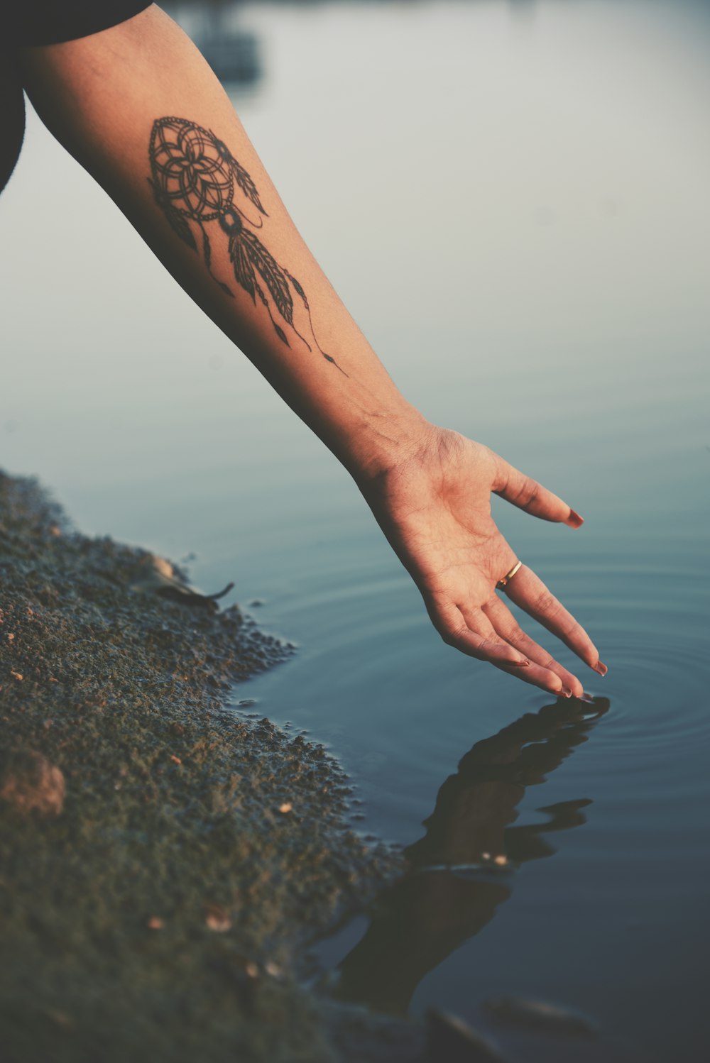 person hand reaching calm water with dreamcatcher tattoo