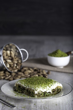 A plate holds a dessert with a layer of white cream topped with finely chopped green pistachios. In the background, a metal container lies spilled with whole pistachios spreading out onto the surface, beside a cup filled with more ground pistachios.