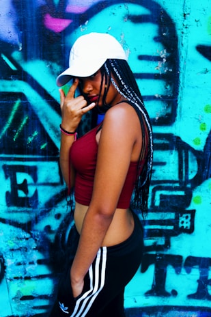 A person with braided hair wearing a white cap and a red sleeveless top poses with a hand gesture in front of a vibrant graffiti-covered wall. The background is predominantly blue with hints of pink and black abstract shapes.