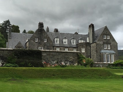 A large stone mansion with multiple chimneys and dormer windows, set against a backdrop of overcast skies. The foreground features manicured lawns and a variety of shrubs and flowers, with lush greenery surrounding the property.