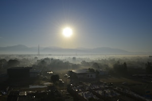 A cityscape during sunrise with the sun shining brightly above the horizon. Mist shrouds the buildings and trees, creating a dreamy atmosphere. Mountains can be seen in the background with a clear blue sky overhead.