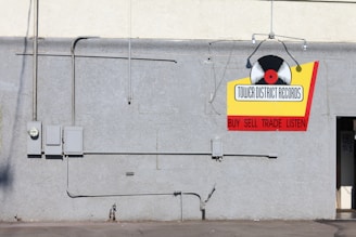 A gray concrete wall with several electrical boxes and conduits. A colorful sign hangs on the wall with a vinyl record graphic and the text 'Tower District Records' along with the words 'Buy Sell Trade Listen' in red. There is a small, partially visible door on the right side.