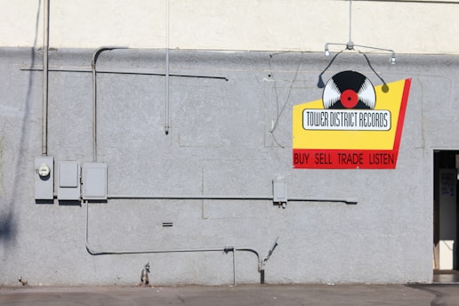 A gray concrete wall with several electrical boxes and conduits. A colorful sign hangs on the wall with a vinyl record graphic and the text 'Tower District Records' along with the words 'Buy Sell Trade Listen' in red. There is a small, partially visible door on the right side.