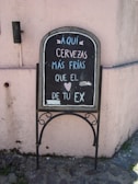 A chalkboard sign placed against a light pink stucco wall, featuring a humorous message in Spanish about cold beers being colder than an ex. It is written in white and light blue chalk, with a small heart symbol included in the text.