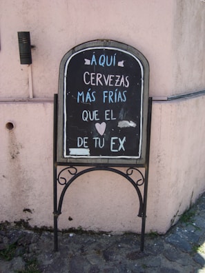 A chalkboard sign placed against a light pink stucco wall, featuring a humorous message in Spanish about cold beers being colder than an ex. It is written in white and light blue chalk, with a small heart symbol included in the text.