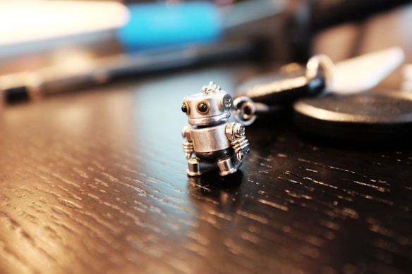 A small silver robot figurine is placed on a wooden table, surrounded by blurred objects in the background, including a pen and various metallic items. The robot has a whimsical and detailed design with mechanical features.