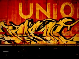 A section of a train car features vibrant graffiti with bold, flowing letters painted predominantly in black and yellow tones. The word 'UNION' appears in large capital letters above the graffiti. The overall surface looks well-worn, conveying a gritty urban atmosphere.