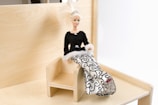 A fashion doll with blonde hair and a tiara is seated on a small wooden chair. She is dressed in a stylish gown with a black top and white skirt featuring intricate black patterns. The setting is minimalistic, with a light wooden background.