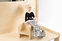 A fashion doll with blonde hair and a tiara is seated on a small wooden chair. She is dressed in a stylish gown with a black top and white skirt featuring intricate black patterns. The setting is minimalistic, with a light wooden background.