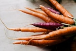 A bundle of fresh carrots with dirt on their surface, including one vibrant purple carrot among the orange ones, placed on a light wooden surface.