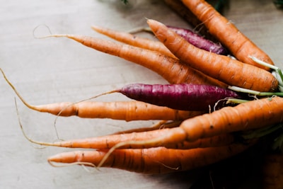 A bundle of fresh carrots with dirt on their surface, including one vibrant purple carrot among the orange ones, placed on a light wooden surface.