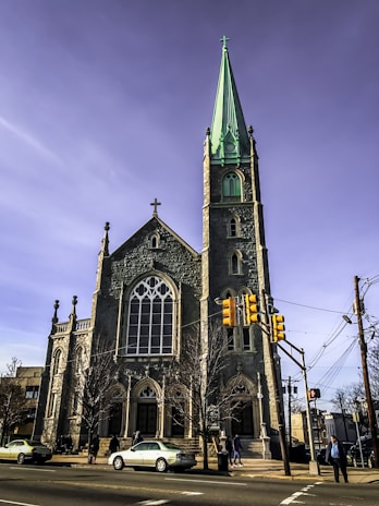 A large stone church with a tall, green spire stands prominently at a street corner. The building features gothic architecture with arched windows and a cross on top. Several cars are parked in front, and a few people are walking nearby. A traffic light and some power lines are visible in the foreground under a clear blue sky.
