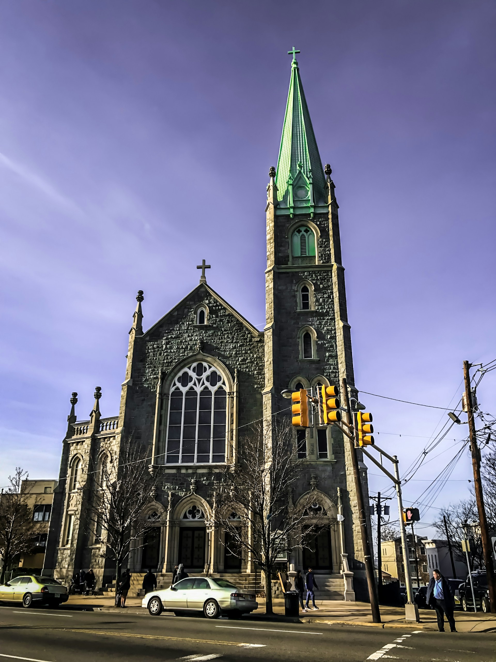 A large stone church with a tall, green spire stands prominently at a street corner. The building features gothic architecture with arched windows and a cross on top. Several cars are parked in front, and a few people are walking nearby. A traffic light and some power lines are visible in the foreground under a clear blue sky.