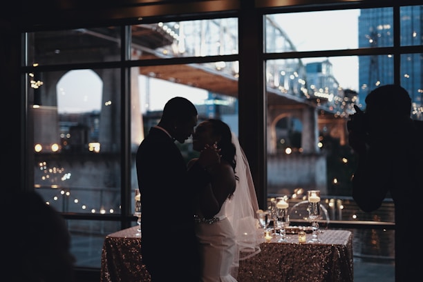 A silhouetted couple is dancing in an elegant room with large windows showcasing a softly lit bridge outside. The room is decorated with a sequined tablecloth and candles creating a romantic ambiance.