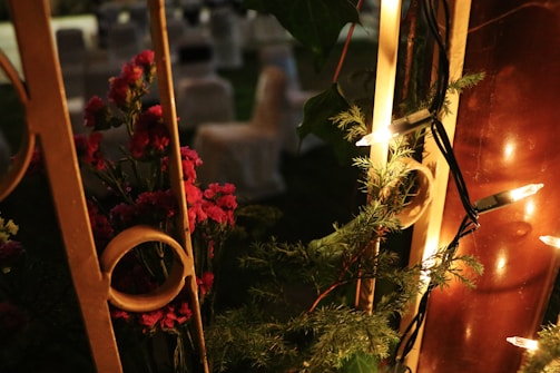 An intimate scene with warm lighting showcases vibrant red flowers intertwined with green foliage. Soft yellow glowing string lights are draped around a wooden structure, creating a cozy, inviting atmosphere. In the background, blurred outlines of chairs suggest a garden setting.