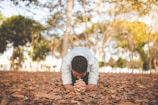 A person is kneeling on a bed of fallen leaves in an outdoor park, with trees in the background. They have their head bowed and hands clasped together in a gesture of contemplation or prayerful reflection. The sunlight filters through the tree branches, creating a warm, serene atmosphere.