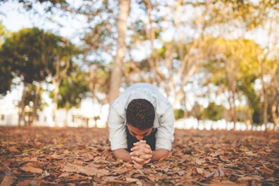A person is kneeling on a bed of fallen leaves in an outdoor park, with trees in the background. They have their head bowed and hands clasped together in a gesture of contemplation or prayerful reflection. The sunlight filters through the tree branches, creating a warm, serene atmosphere.