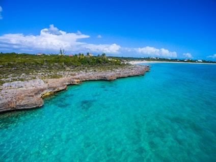 A crystal clear turquoise ocean hugs a rocky shoreline under a vibrant blue sky dotted with white clouds. Sparse vegetation grows on the land, leading to a distant sandy beach. The vivid colors and natural scenery create a serene and inviting atmosphere.