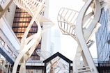 Tall, twisting architectural structures made of metal are prominently featured amid urban high-rises. The structures resemble open sails or abstract helixes, adding a modern and artistic element to the urban environment. The buildings surrounding the structures have reflective glass surfaces, lending a sleek and contemporary appearance.