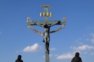 A large crucifix stands against a clear blue sky, featuring a statue of Jesus Christ with outstretched arms. Surrounding the cross are gold-colored Hebrew letters that form a semicircle. Two figures dressed in dark robes stand at the base, gazing upward.