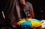 Close-up of hands cutting a beautifully decorated birthday cake surrounded by smiling guests.