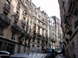 Narrow street lined with classic Parisian-style apartment buildings featuring ornate iron balconies and window boxes with flowers. Cars are parked along the sides, and the architecture reflects an elegant, historical atmosphere.
