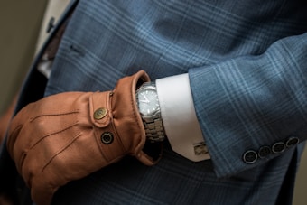 A close-up of a person wearing a sophisticated plaid blue suit with noticeable button details on the sleeve. The person is also wearing a brown leather glove, and underneath the glove, a silver wristwatch is visible. The watch adds a touch of elegance to the ensemble.