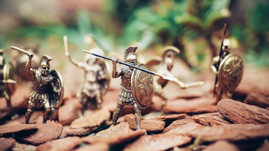 Miniature figurines of ancient warriors are posed in an action scene. They are equipped with shields, spears, and helmets, resembling historical soldiers. The background is blurred, giving focus to the detailed textures and metallic sheen of the figures.
