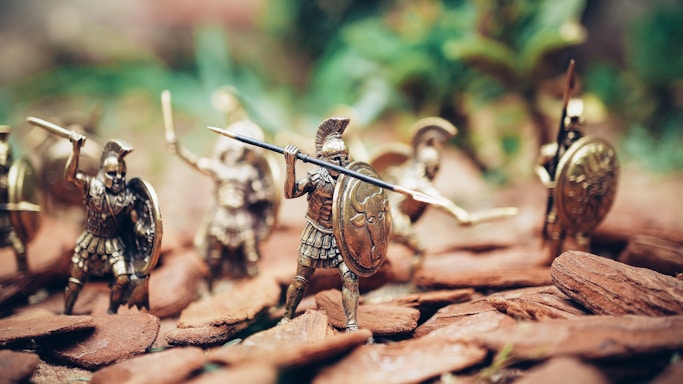Miniature figurines of ancient warriors are posed in an action scene. They are equipped with shields, spears, and helmets, resembling historical soldiers. The background is blurred, giving focus to the detailed textures and metallic sheen of the figures.