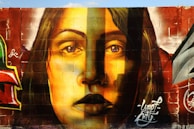A mural painted on a brick wall, featuring the realistic portrait of a woman with long hair. The artwork uses a vibrant color palette, primarily in shades of yellow, brown, and red. The detailed shading and expression on her face create a striking visual impact. Surrounding elements include graffiti art and signatures in various colors, adding an urban feel.