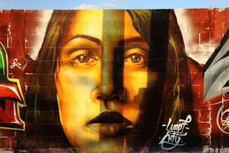 A mural painted on a brick wall, featuring the realistic portrait of a woman with long hair. The artwork uses a vibrant color palette, primarily in shades of yellow, brown, and red. The detailed shading and expression on her face create a striking visual impact. Surrounding elements include graffiti art and signatures in various colors, adding an urban feel.