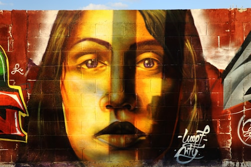 A mural painted on a brick wall, featuring the realistic portrait of a woman with long hair. The artwork uses a vibrant color palette, primarily in shades of yellow, brown, and red. The detailed shading and expression on her face create a striking visual impact. Surrounding elements include graffiti art and signatures in various colors, adding an urban feel.