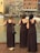 Three women in brown dresses stand on a wooden deck overlooking a body of water, with their backs facing the camera. They are near a railing and appear to be at a waterfront area with buildings in the background. One of the women holds a bouquet of flowers, and a pair of shoes is placed on the ground next to one woman.