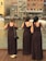 Three women in brown dresses stand on a wooden deck overlooking a body of water, with their backs facing the camera. They are near a railing and appear to be at a waterfront area with buildings in the background. One of the women holds a bouquet of flowers, and a pair of shoes is placed on the ground next to one woman.