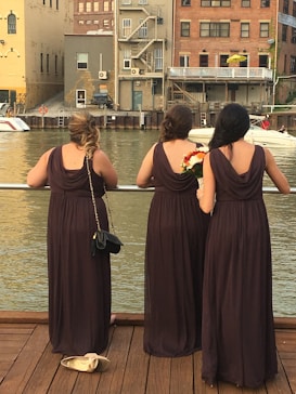Three women in brown dresses stand on a wooden deck overlooking a body of water, with their backs facing the camera. They are near a railing and appear to be at a waterfront area with buildings in the background. One of the women holds a bouquet of flowers, and a pair of shoes is placed on the ground next to one woman.