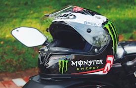 A motorcycle helmet with a clear visor and prominent Monster Energy branding is positioned on a parked motorcycle. The logo is green, white, and red on the side of the black helmet. In the background, there is a grassy area with some fallen leaves.