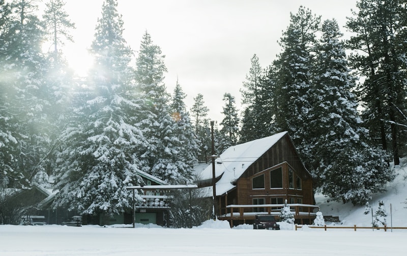 A cozy wooden cabin nestled in a snowy forest with tall pine trees surrounding it. The sun is peeking through the trees, casting a gentle light on the snow-covered landscape. A vehicle is parked near the cabin, and the scene is enveloped in a serene and peaceful atmosphere.