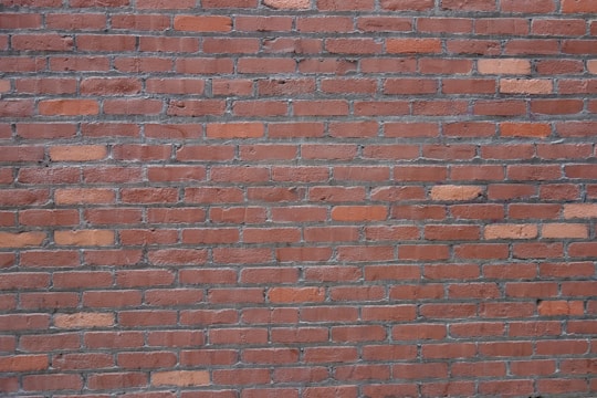 A surface composed of evenly stacked red bricks held together by gray mortar. The bricks have a uniform rectangular shape and display various shades of red and brown tones. The texture is rough, and there are slight inconsistencies in brick placement, adding character to the wall.