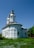 A white church with a simple architectural design featuring two towers topped with crosses. It is surrounded by greenery and wildflowers, under a clear blue sky.