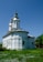 A white church with a simple architectural design featuring two towers topped with crosses. It is surrounded by greenery and wildflowers, under a clear blue sky.