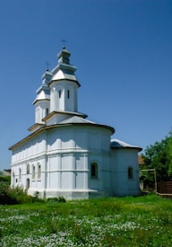 A white church with a simple architectural design featuring two towers topped with crosses. It is surrounded by greenery and wildflowers, under a clear blue sky.