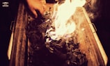 A hand is adjusting pieces of charcoal that are ablaze in a metal grill or container. The glowing flames create a warm light, contrasting with the dark, charred wood.