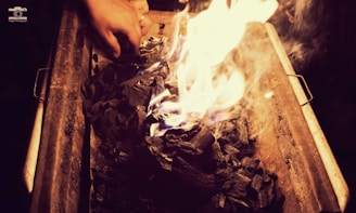 A hand is adjusting pieces of charcoal that are ablaze in a metal grill or container. The glowing flames create a warm light, contrasting with the dark, charred wood.
