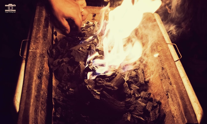 A hand is adjusting pieces of charcoal that are ablaze in a metal grill or container. The glowing flames create a warm light, contrasting with the dark, charred wood.