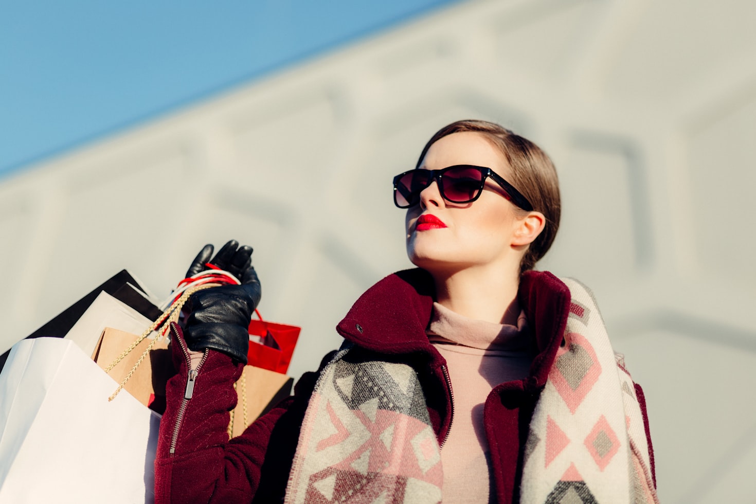 Woman wearing sunglasses, burgundy coat, and large scarf holds a bunch of shopping bags while looking up. Remember that a global work and travel abroad experience usually does not mean making a ton of money!
