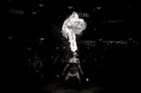 A performer is seen in a dramatic act of fire breathing, releasing a large plume of fire upwards. The scene is set against a dark background, emphasizing the intensity of the flames and drawing attention to the performer. Surrounding the performer are blurred silhouettes of an audience, creating a sense of focus and spotlight on the act.