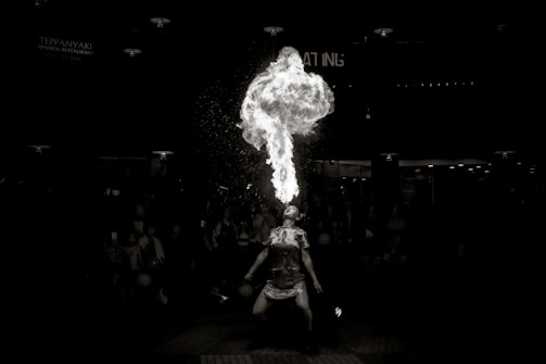 A performer is seen in a dramatic act of fire breathing, releasing a large plume of fire upwards. The scene is set against a dark background, emphasizing the intensity of the flames and drawing attention to the performer. Surrounding the performer are blurred silhouettes of an audience, creating a sense of focus and spotlight on the act.
