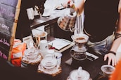 A barista is preparing coffee using a pour-over method with a kettle, pouring hot water over ground coffee in a filter over a glass carafe. Various coffee-related items such as jars of coffee beans, paper cups, and a menu board with chalk writing are visible on the counter.