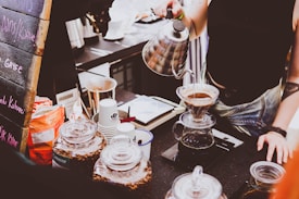 A barista is preparing coffee using a pour-over method with a kettle, pouring hot water over ground coffee in a filter over a glass carafe. Various coffee-related items such as jars of coffee beans, paper cups, and a menu board with chalk writing are visible on the counter.